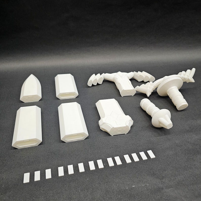 May include: A collection of white 3D-printed components, including geometric shapes, handles, and a cylindrical piece with a knob. The parts are arranged on a dark surface, suggesting a kit or set of modular elements.