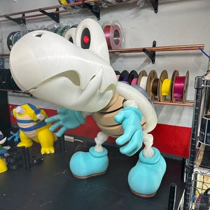 May include: A white and blue 3D printed figurine of a skeletal dinosaur with a large head and a single red eye. The figurine is standing on a black surface.
