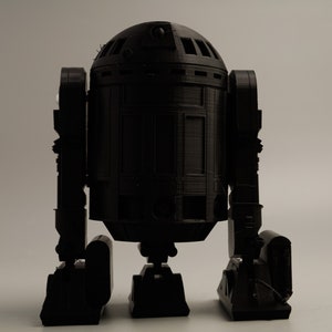 Op de afbeelding: Een zwart 3D-geprint model van een droid met een afgeronde kop en rechthoekig lichaam. De droid heeft twee benen en twee armen en staat op een wit oppervlak. Het model is een replica van een populair sciencefiction-personage.