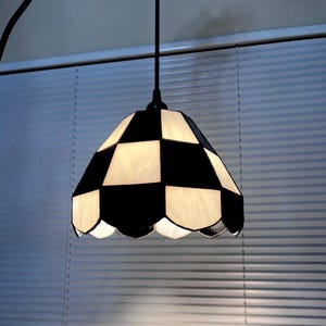 May include: A hanging pendant lamp with a black and white checkered shade. The shade has a scalloped edge and is made of glass or a similar material. The lamp is suspended from a black cord and is against a window with blinds.