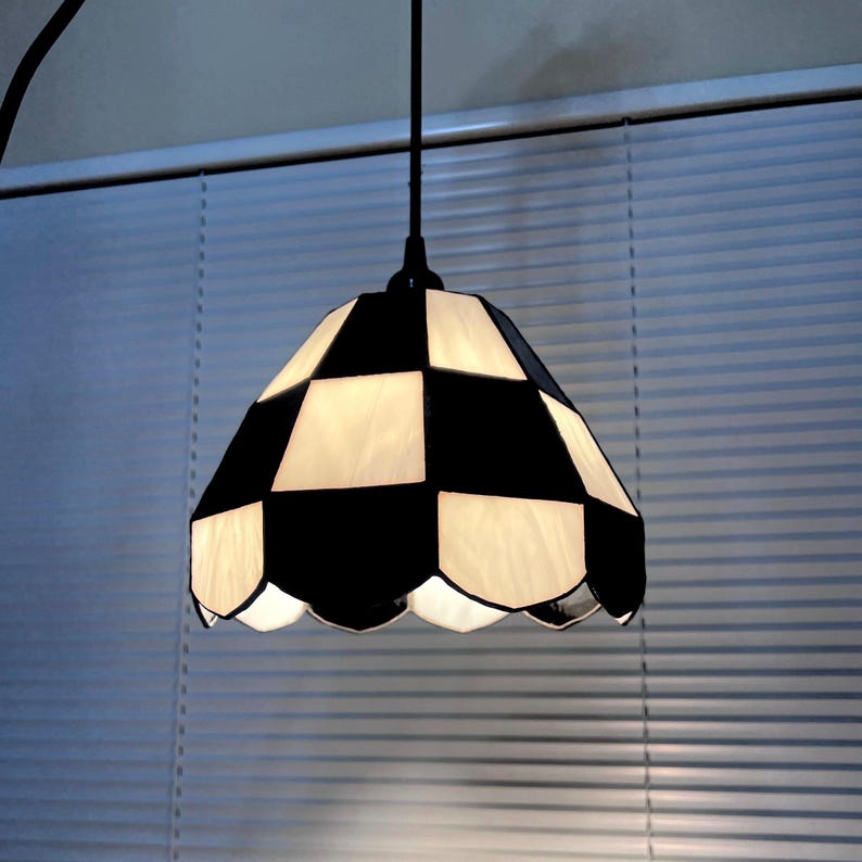 May include: A hanging pendant lamp with a black and white checkered shade. The shade has a scalloped edge and is made of glass or a similar material. The lamp is suspended from a black cord and is against a window with blinds.