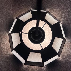 Peut inclure: Vue de dessus d'un luminaire suspendu g&eacute;om&eacute;trique noir et blanc. L'abat-jour est octogonal avec des panneaux noirs et blancs altern&eacute;s. La source de lumi&egrave;re est visible au centre, d&eacute;gageant une lueur chaude.