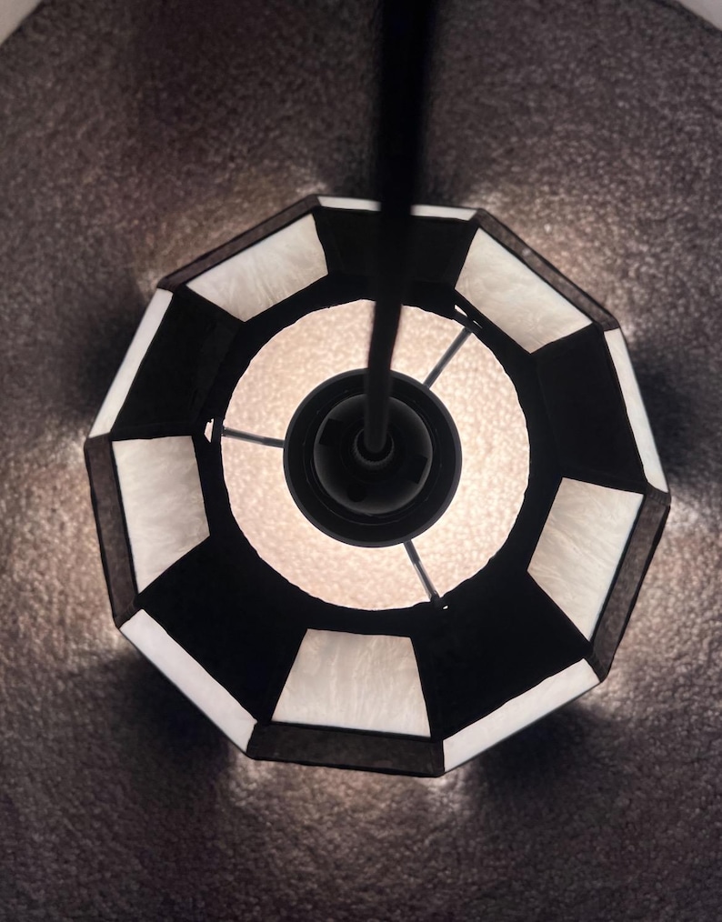 Peut inclure: Vue de dessus d'un luminaire suspendu g&eacute;om&eacute;trique noir et blanc. L'abat-jour est octogonal avec des panneaux noirs et blancs altern&eacute;s. La source de lumi&egrave;re est visible au centre, d&eacute;gageant une lueur chaude.