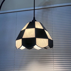 May include: A hanging pendant light with a black and white checkered lampshade. The shade has a scalloped edge and is suspended from a black cord. The light fixture is designed to provide ambient lighting.