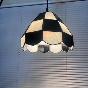 Peut inclure: Une suspension luminaire avec un abat-jour &agrave; carreaux noirs et blancs. L'abat-jour a un bord festonn&eacute; et un cordon noir. La lumi&egrave;re est allum&eacute;e, &eacute;clairant l'int&eacute;rieur.
