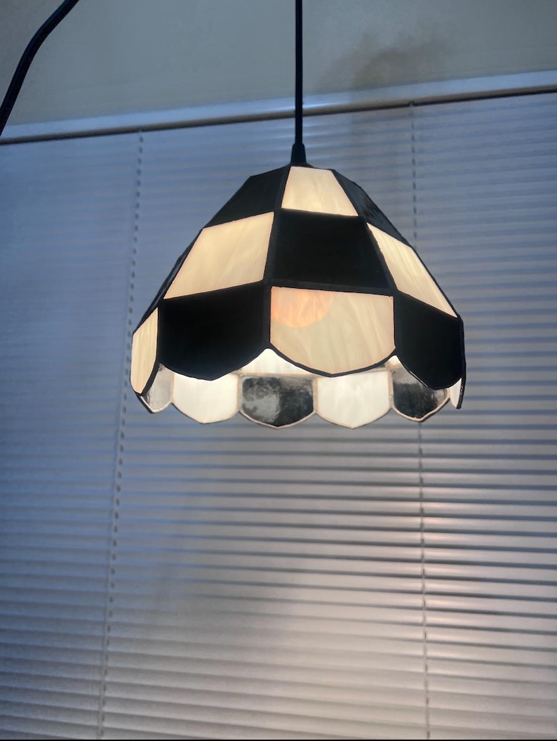 Peut inclure: Une suspension luminaire avec un abat-jour &agrave; carreaux noirs et blancs. L'abat-jour a un bord festonn&eacute; et un cordon noir. La lumi&egrave;re est allum&eacute;e, &eacute;clairant l'int&eacute;rieur.