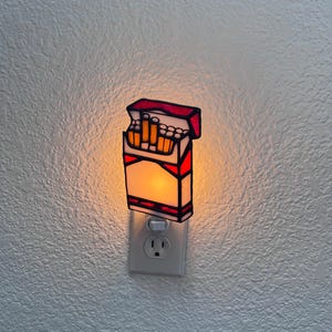 May include: A decorative night light shaped like a pack of cigarettes. The light has a white body with red accents and a red top. The cigarettes inside are orange. The night light is plugged into a standard wall outlet.