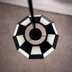 May include: Overhead view of a black and white geometric pendant light fixture. The lamp shade has a repeating pattern of black and white trapezoids. The light fixture is suspended from a black rod.