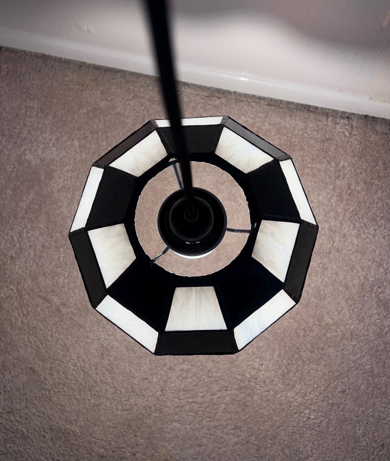 May include: Overhead view of a black and white geometric pendant light fixture. The lamp shade has a repeating pattern of black and white trapezoids. The light fixture is suspended from a black rod.