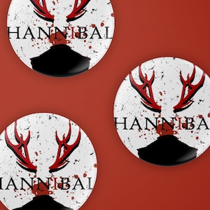 May include: Three white round buttons with a black silhouette of a head with red antlers and blood splatters. The text "HANNIBAL" is printed in black on each button.