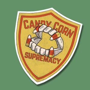 Könnte beinhalten: Ein gelber und roter schildförmiger Aufkleber mit dem Text "Candy Corn Supremacy" und einer Grafik von einem Gebiss mit Candy Corn anstelle der Zähne.