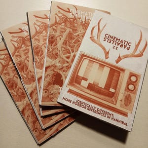 Op de afbeelding: Een stapel bruin-witte boekjes getiteld "Cinematic Parallels II". De omslag toont een vintage televisie met een testpatroon en geweien. De boekjes hebben een sepia-achtige uitstraling en de tekst "Identiek anders: Meer horrorreferenties in Hannibal."
