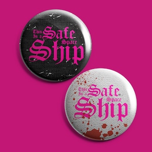 Puede incluir: Dos botones redondos con el texto "This is a Safe Space Ship" en letras góticas rosas. Un botón tiene un fondo negro con una textura desgastada. El otro botón tiene un fondo blanco con salpicaduras rojas.