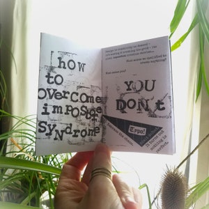 May include: A small, black and white book with the text "How to Overcome Imposter Syndrome" on the left page and "You Don't" on the right page. The text is in a bold, blocky font. The book is open and being held by a hand.