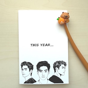 May include: A white card with the text "THIS YEAR..." and illustrations of three men in black and white. A brown pen with a bear-shaped topper is on the right side of the card.
