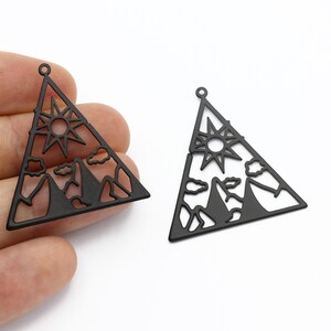 May include: Two black metal triangle charms featuring a mountain and sun design. The charms are cut out and have a hole at the top for hanging.
