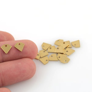 May include: A close-up of a pile of small, gold-colored metal charms shaped like a diamond with a hole in the top. Two of the charms are held in a hand.