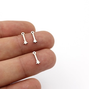 Op de afbeelding: Drie zilveren stervormige bedels met een lus aan de bovenkant en een rechte lijn die van de lus uitstrekt. De bedels bevinden zich op een vinger.