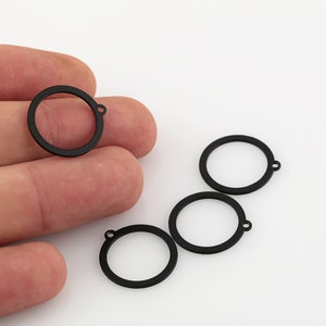 May include: Four black metal circle charms with a jump ring attached to each. The charms are on a white surface.