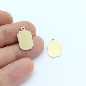May include: Two small, gold-colored rectangular pendants with a loop at the top. The pendants have a geometric, octagonal shape. One pendant is held in a person's hand, while the other is on a white surface.
