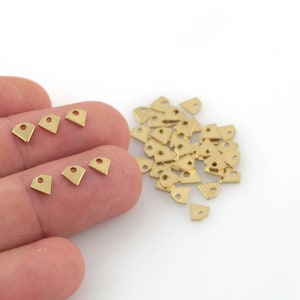 Könnte beinhalten: Ein Haufen kleiner, goldfarbener Metall-Dreiecksanhänger mit einem Loch in der Mitte. Die Anhänger sind auf einer weißen Oberfläche verstreut. Eine Hand hält ein paar der Anhänger.