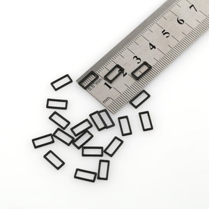 May include: A close-up of a metal ruler with black markings and numbers, measuring in millimeters, next to a scattering of small black rectangular shapes on a white surface.