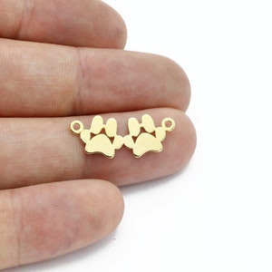 May include: A gold-colored charm featuring two paw prints connected by small loops. The charm is designed to be used in jewelry making, such as bracelets or necklaces. The paw prints are detailed and have a polished finish.