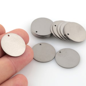 May include: A close-up of a pile of round, silver metal discs with a small hole in the center of each disc. The discs are smooth and have a slightly brushed finish.