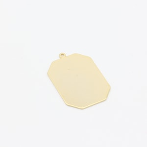 May include: A gold-colored, rectangular pendant with a small loop at the top. The pendant has a smooth, polished surface and a geometric shape with slightly rounded corners. The background is plain white.