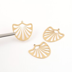 May include: Three gold-colored, fan-shaped pendants with a cut-out design. One pendant is held by tweezers. The pendants have a small loop at the top for attaching to jewelry. The pendants are on a white background.