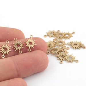 May include: A close-up of a hand holding gold-toned metal starburst charms with two jump rings on each charm. The charms are scattered on a white surface.