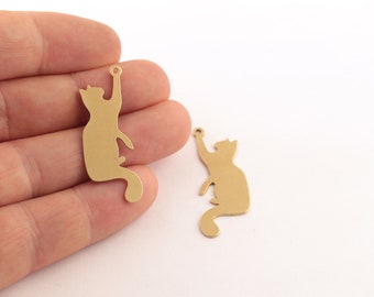 Puede incluir: Dos dijes de gato de metal dorado, cada uno con un agujero en la parte superior para colgar. Los gatos están en una pose juguetona, con las patas levantadas en el aire.