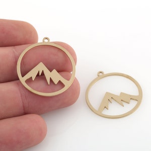 May include: Two gold-colored metal charms in the shape of a circle with a mountain silhouette cut out.
