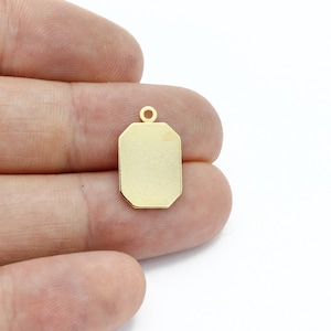 May include: A gold-colored rectangular pendant with a small loop at the top. The pendant has a smooth, polished surface and a geometric, octagonal shape. The pendant is held in a person's hand against a white background.