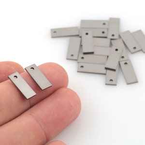 May include: A close-up of two small, silver, rectangular metal tags with a hole in the top. The tags are held in a hand. There are several more tags in the background.
