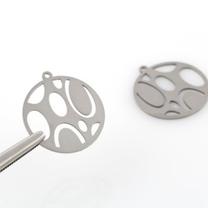 May include: Two silver metal charms with a circular shape and a cut-out design of abstract shapes. The charms have a small loop at the top for attaching to jewelry.