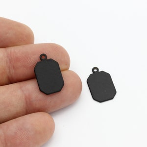 May include: Two black metal pendant charms with a rectangular shape and a slightly rounded edge. The charms have a small loop at the top for attaching to a chain or cord.