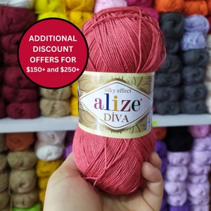 Op de afbeelding: Een bol Alize Diva silky effect garen in een koraalkleur. Het etiket toont de merknaam en productdetails. Het garen wordt in een hand gehouden, met planken met verschillende garenkleuren op de achtergrond.