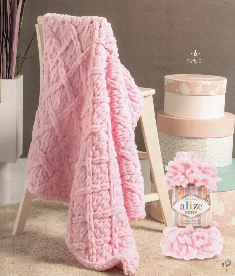 Puede incluir: Una manta rosa p&aacute;lido de punto grueso con un patr&oacute;n geom&eacute;trico, sobre una peque&ntilde;a silla de madera. Junto a la silla hay tres cajas redondas decorativas y un paquete de hilo Alize Puffy.