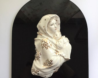May include: A ceramic wall plaque depicting a mother and child. The figures are white with gold floral detailing, set against a glossy black arched background. The plaque is mounted with a silver hook.