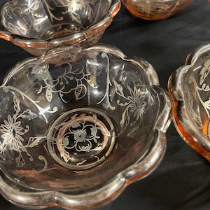 Könnte beinhalten: Eine Sammlung rosa Glasschalen mit silbernen Blumendesigns. Die Schalen haben einen gewellten Rand und einen silbernen Rand. Das Blumenmuster umfasst Blumen und Blätter. Die Schalen sind auf einer dunklen Oberfläche angeordnet.