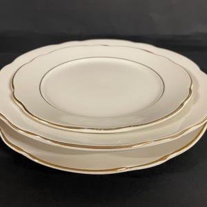 Könnte beinhalten: Ein Stapel aus drei weißen Keramikplatten mit gewellten Rändern und goldenen Verzierungen. Die Teller sind unterschiedlich groß, der kleinste Teller liegt oben. Die Teller stehen auf einer schwarzen Oberfläche.