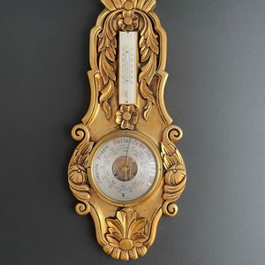 May include: A vintage gold-coloured barometer and thermometer combination. The ornate frame features floral and leaf designs. The thermometer displays a temperature scale. The barometer dial has Italian text, including "Pioggia" and "Variabile".