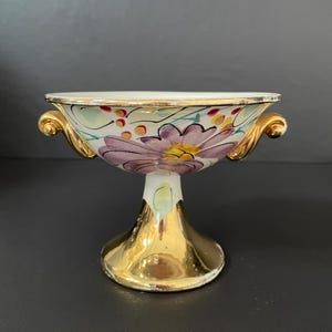 May include: A white ceramic bowl with gold accents and floral designs in shades of purple, yellow, and green. The bowl is on a gold pedestal with a round base.