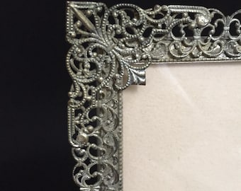 May include: Close-up of a vintage silver-toned picture frame. The frame features intricate filigree detailing along the edges and corners. The frame's design includes floral and scroll patterns, with a light-colored background visible within the frame.