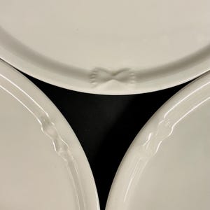 Può includere: Primo piano di tre piatti in ceramica bianca. I piatti hanno un design semplice con un bordo leggermente rialzato. Un piatto presenta un'indentazione a forma di farfalla. I piatti sono disposti su uno sfondo nero.