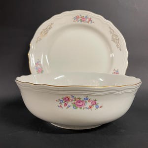 Può includere: Un set di ciotola e piatto in porcellana bianca. La ciotola ha un bordo smerlato e un motivo floreale con fiori rosa, blu e gialli. Il piatto ha anche un bordo smerlato e finiture dorate.