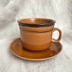 Può includere: Un set di tazza e piattino in ceramica marrone. La tazza ha una forma arrotondata con un manico e una fascia marrone scuro sul bordo. Il piattino è di colore marrone abbinato. Il set è appoggiato su un tessuto di colore neutro.