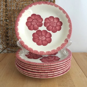 May include: A stack of cream-colored ceramic plates with a floral design. Each plate features three burgundy flowers and a scalloped burgundy border. The plates are stacked on a wooden surface.