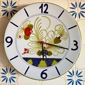 Peut inclure: Horloge murale ronde en céramique blanche avec chiffres et aiguilles noirs. Le cadran présente un motif floral avec des accents rouges, verts et bleus. L'horloge est fixée sur un mur carrelé.